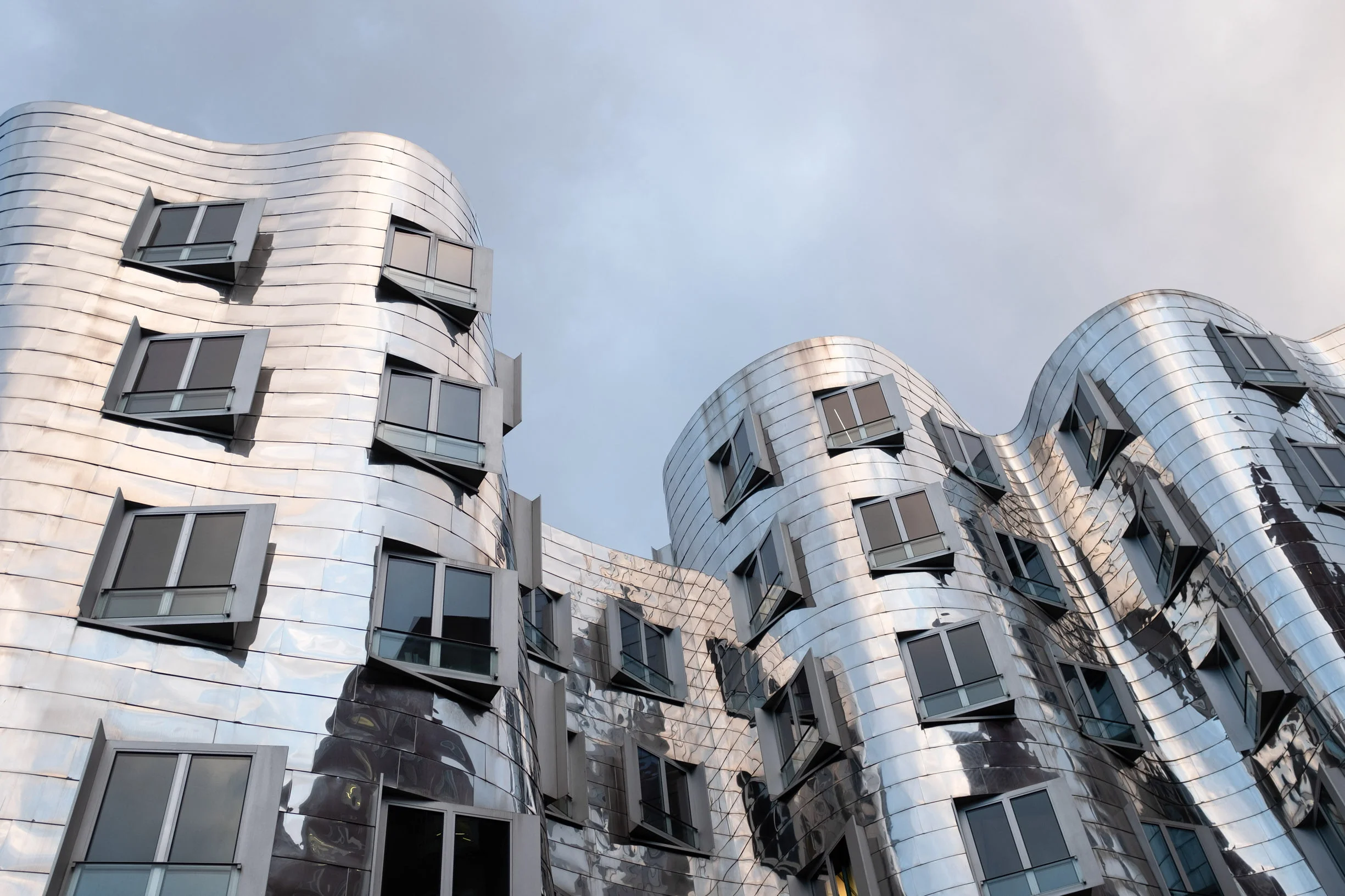 Blick von unten auf Frank Gehry mittleres Gebäude in Düsseldorf. Verspiegelte Fassade und dunkler Himmel. Geschwungene, verchromte Linien.