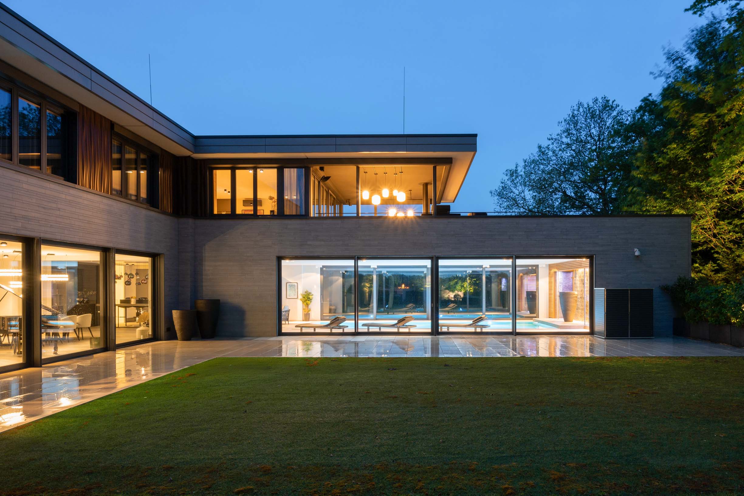 Villa Meerbusch Moderne Villa mit Flachdach, großer Glasfront und beleuchtetem Innenraum in der blauen Stunde, Blick über Rasen und Terrasse.